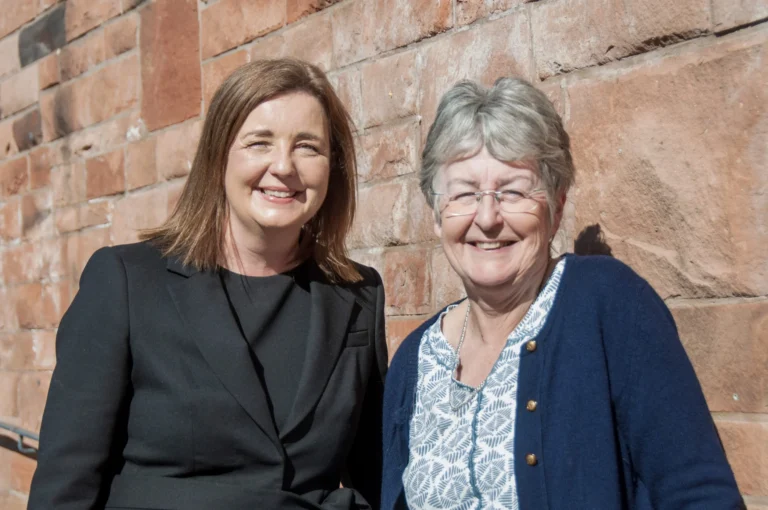 Two women smiling in front of a red brick wall outdoors on a bright day.