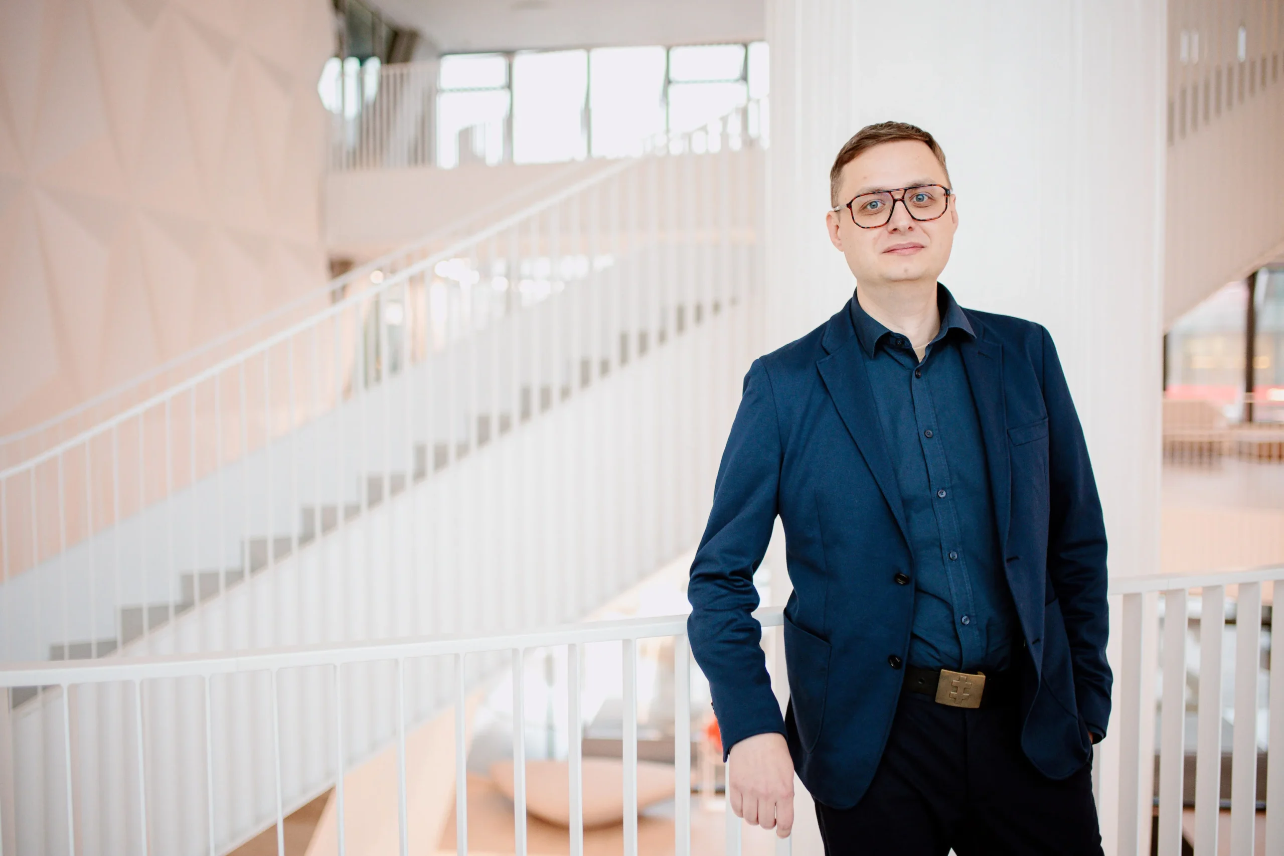 Man wearing glasses and a blue blazer standing in a bright modern office with stairs.