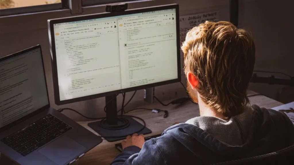 A young man sitting at a desk with a laptop and a large monitor, demonstrating focused digital literacy in a home office setting.