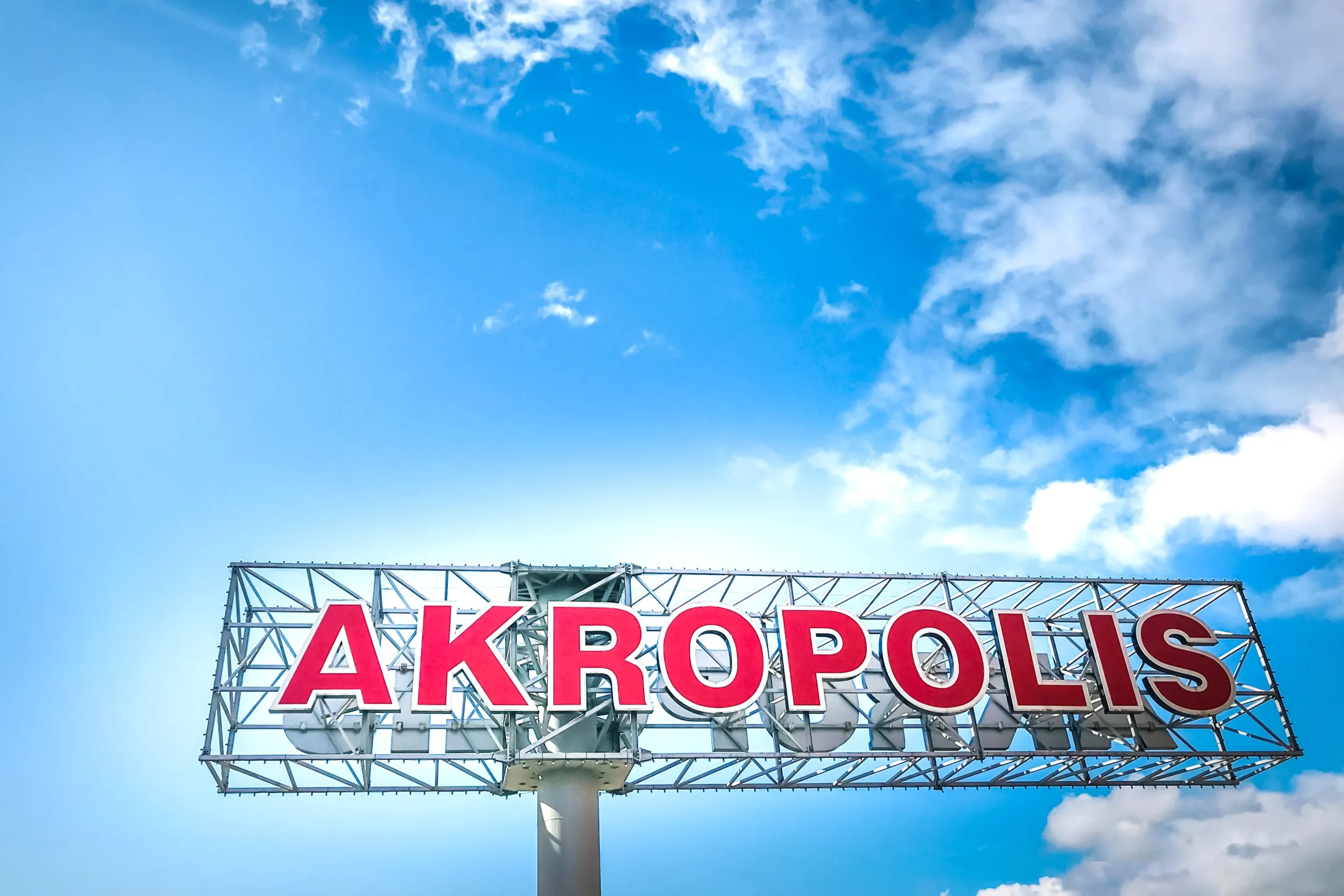 Large red Akropolis sign on a metal framework set against a bright blue cloudy sky.