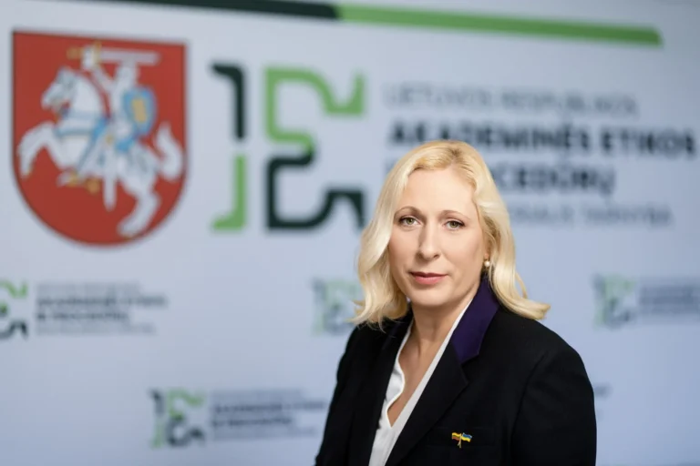 A blonde woman in a black blazer stands before a Lithuanian governmental logo backdrop.