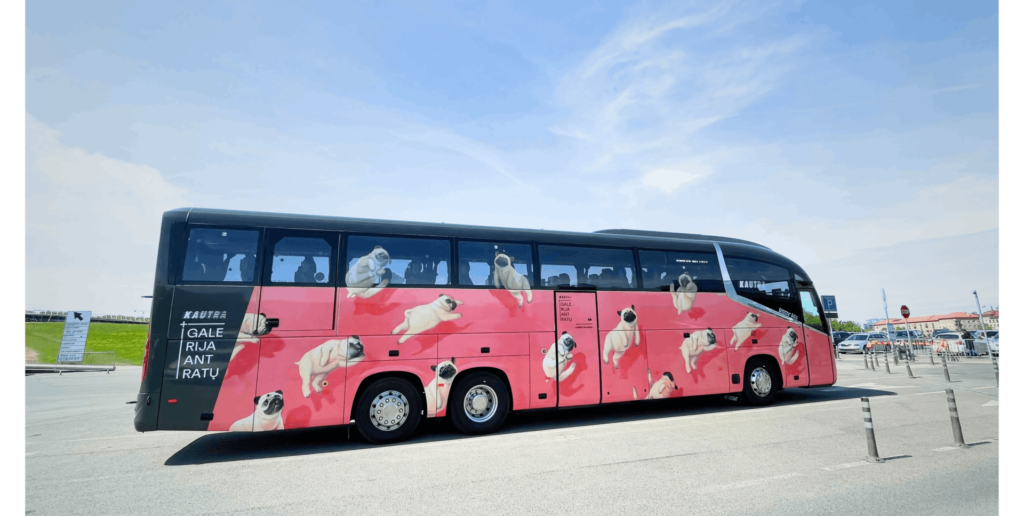 Black coach bus with a pink wrap featuring multiple floating pug dog illustrations parked outdoors.