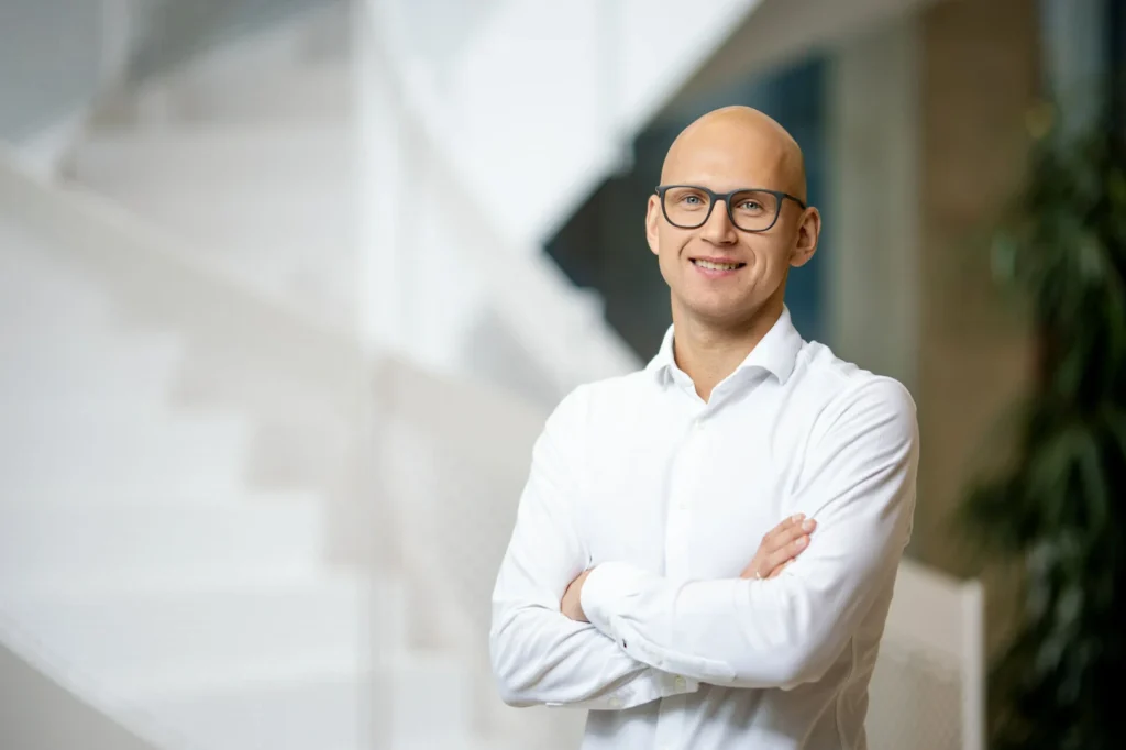 Bald man with glasses and a white shirt smiling with arms crossed in an office.