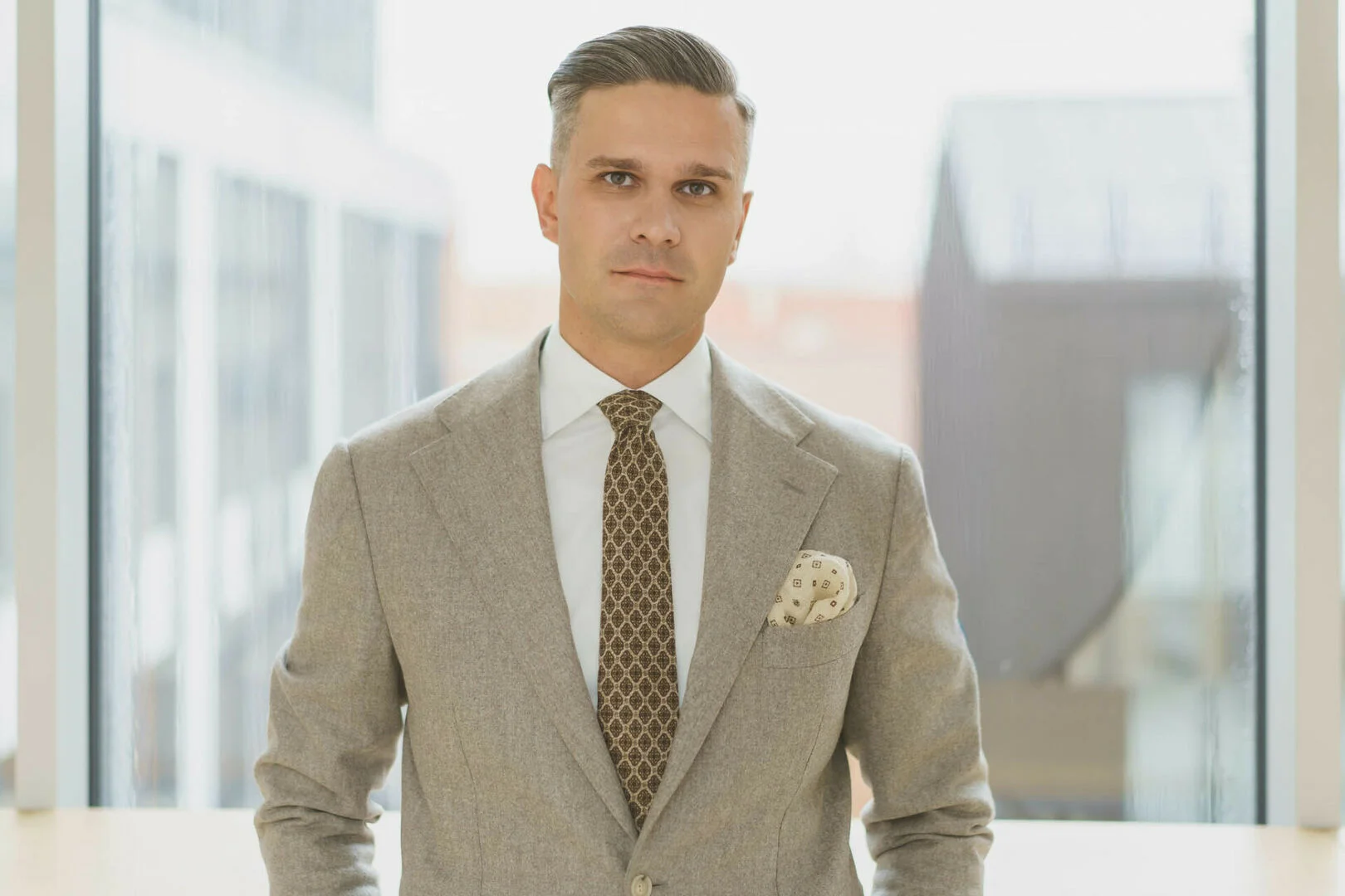 A professional man wearing a tan blazer and patterned tie stands before a bright office window.