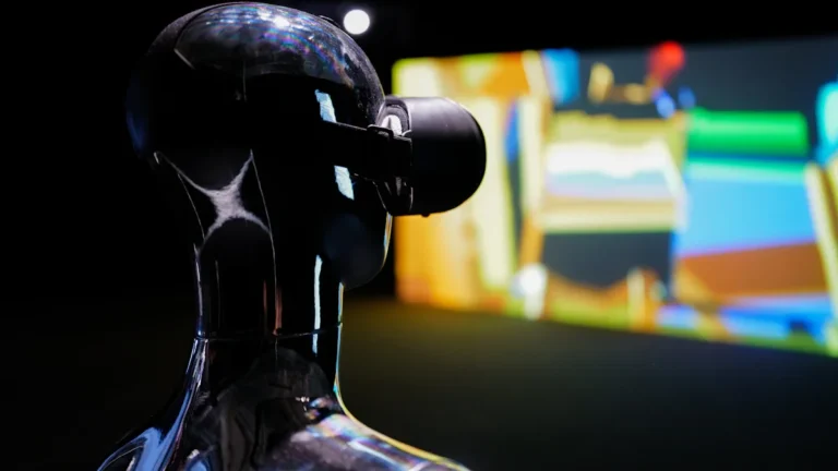 A futuristic humanoid robot bust wearing a VR headset, looking towards a brightly lit screen.