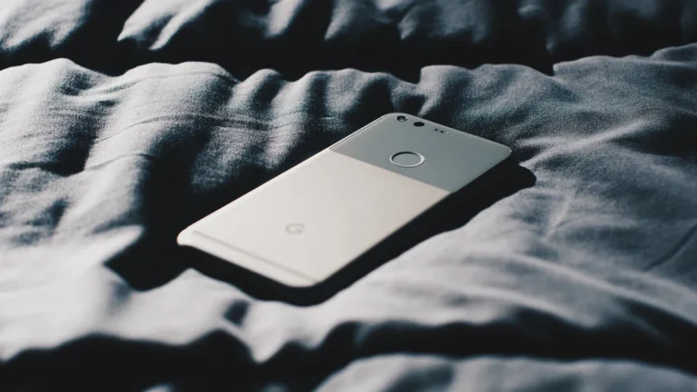 A silver smartphone lies on rumpled grey bedding in a dimly lit, intimate bedroom setting.