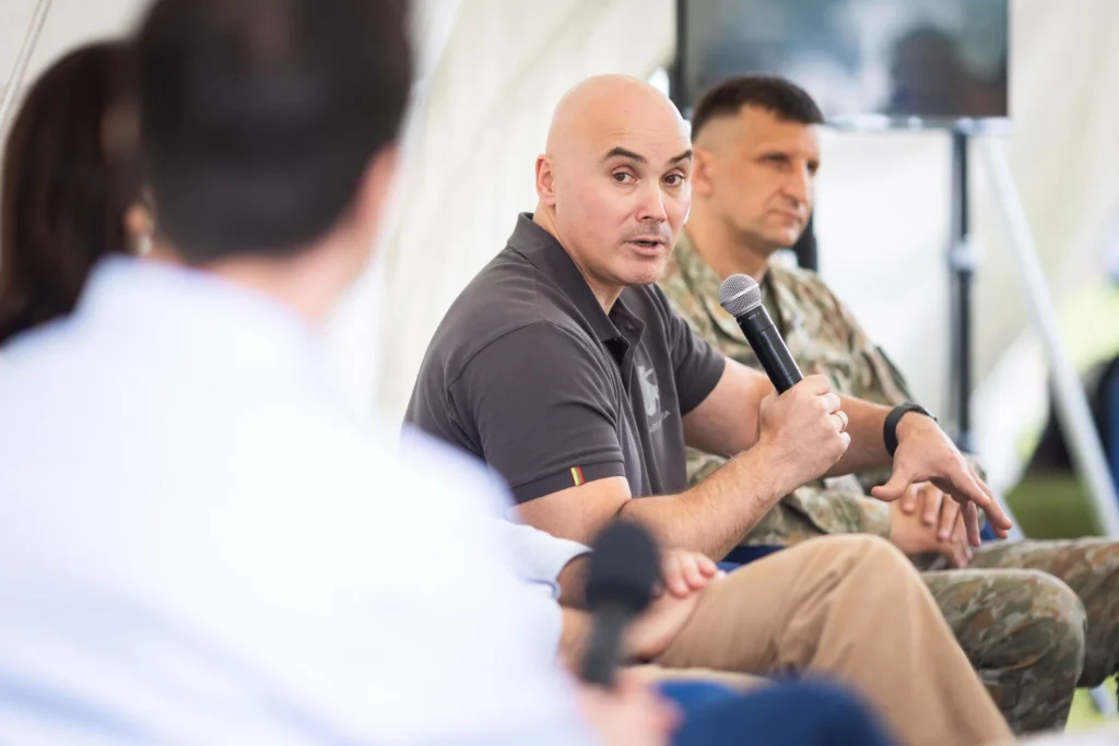 Bald man in grey polo shirt speaks into a microphone sitting beside a soldier in camouflage.