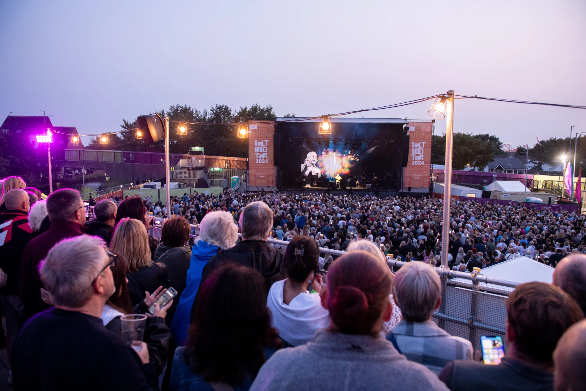 Crowd watching a live concert at an outdoor stage under twilight skies and string lights.
