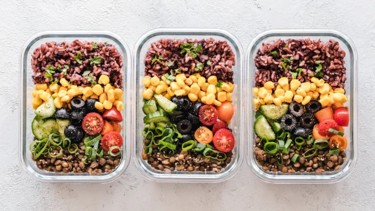 Three glass meal prep containers filled with colorful layers of rice, corn, tomatoes, and lentils.