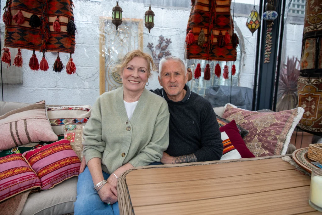 A smiling senior couple sits on a sofa decorated with colorful tasseled rugs and patterned pillows.