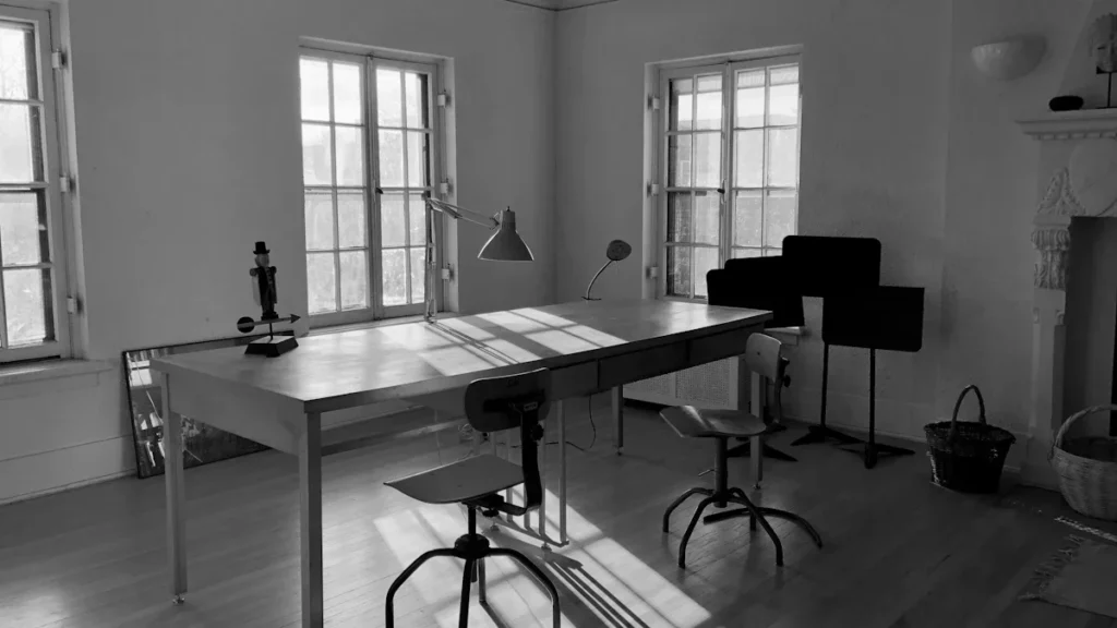 A minimalist, high-ceilinged office space with a large wooden desk, chairs, and natural window lighting.