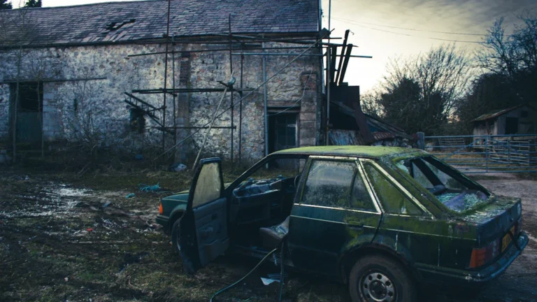 A moss-covered green car sits abandoned and derelict outside a stone building with scaffolding.