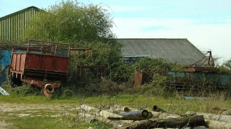 An old, rusty trailer and overgrown weeds near abandoned farm buildings in the English countryside.