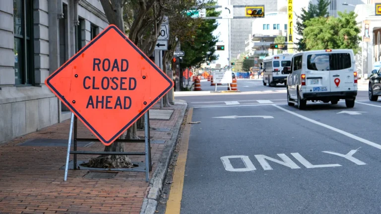 A 'Road Closed Ahead' sign placed on a sidewalk next to a city street with traffic.