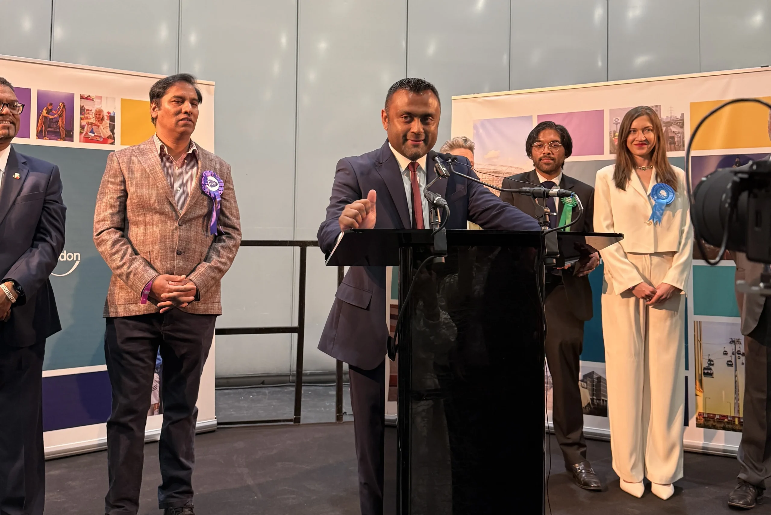 A speaker addresses the audience from a black podium, flanked by candidates wearing colorful rosettes.