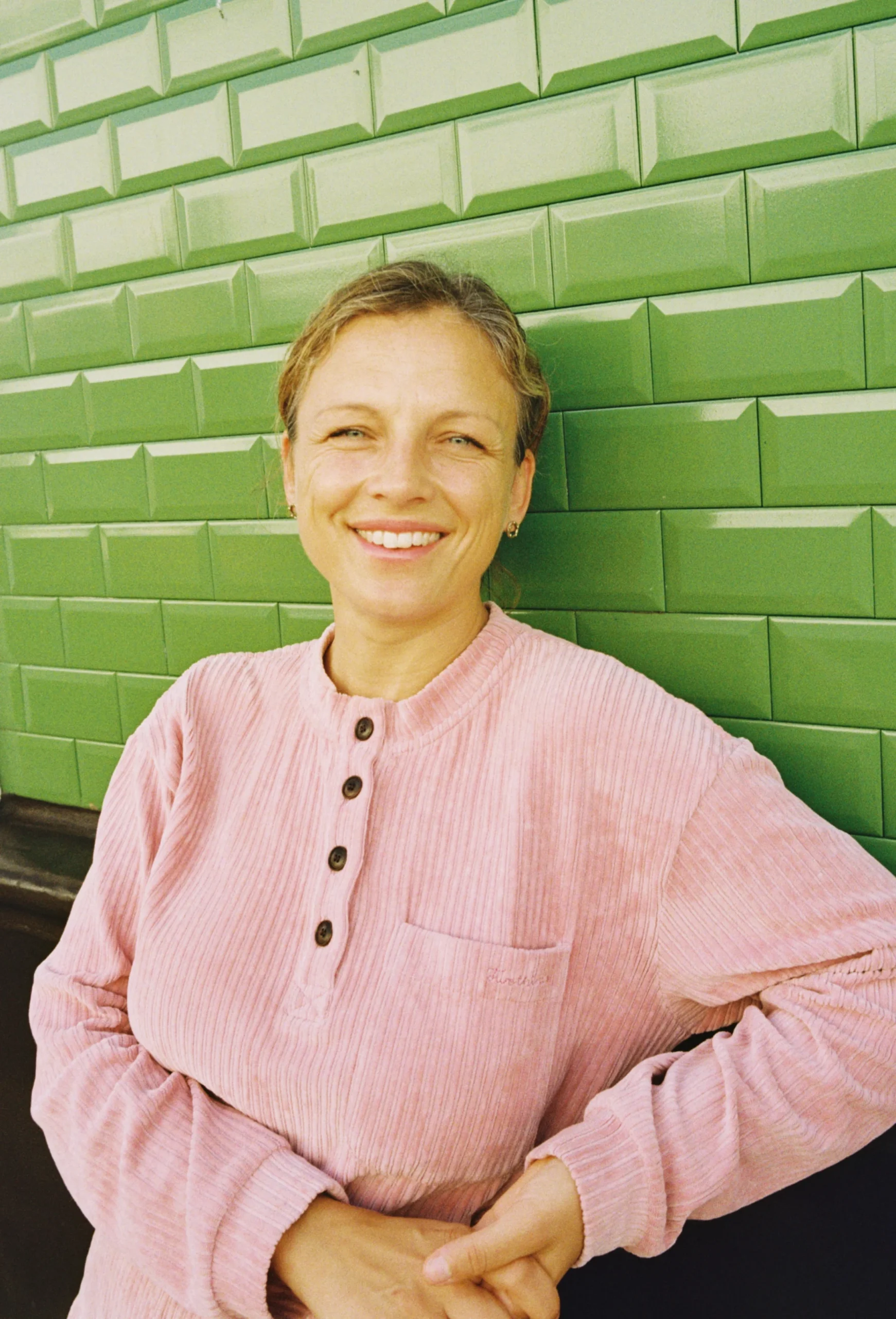 A smiling woman in a pink corduroy shirt poses against a vibrant green tiled wall.