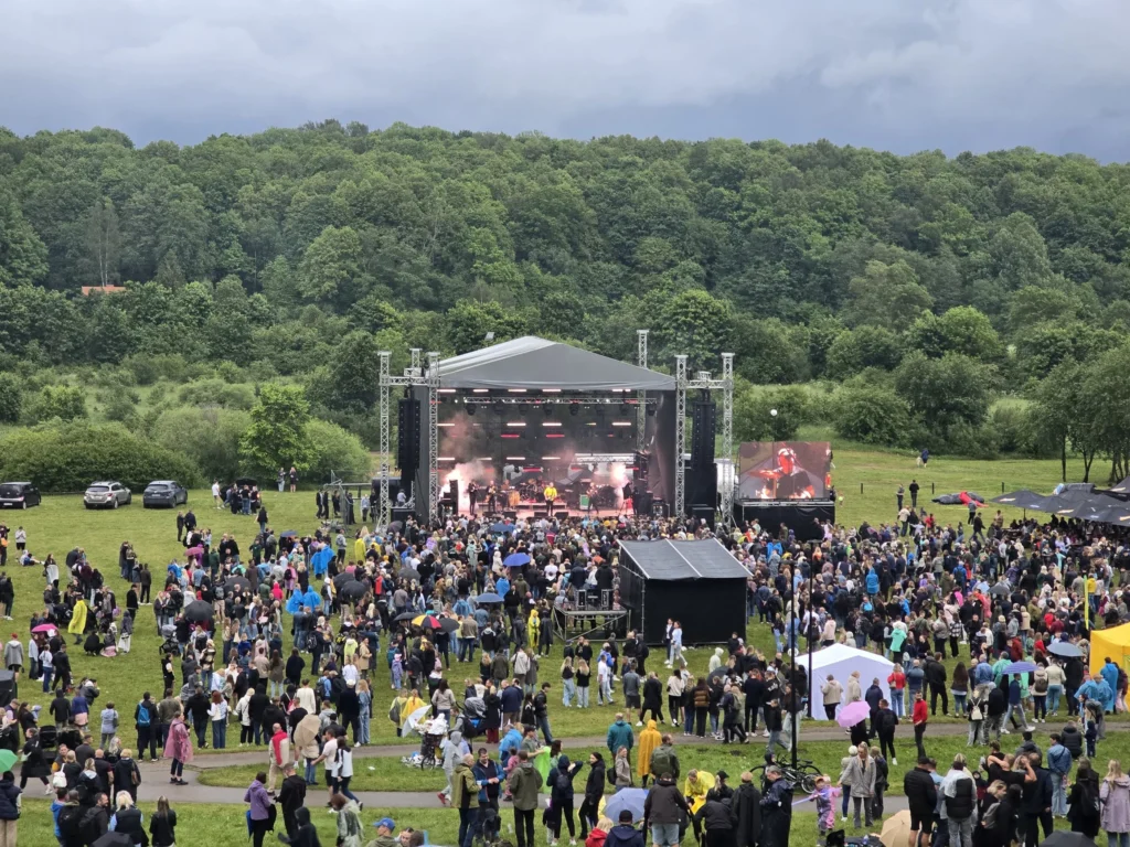 Crowd gathered before an outdoor concert stage with a backdrop of lush green forest hills.