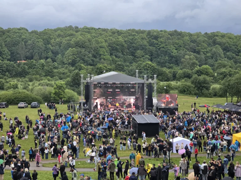 Crowd gathered before an outdoor concert stage with a backdrop of lush green forest hills.