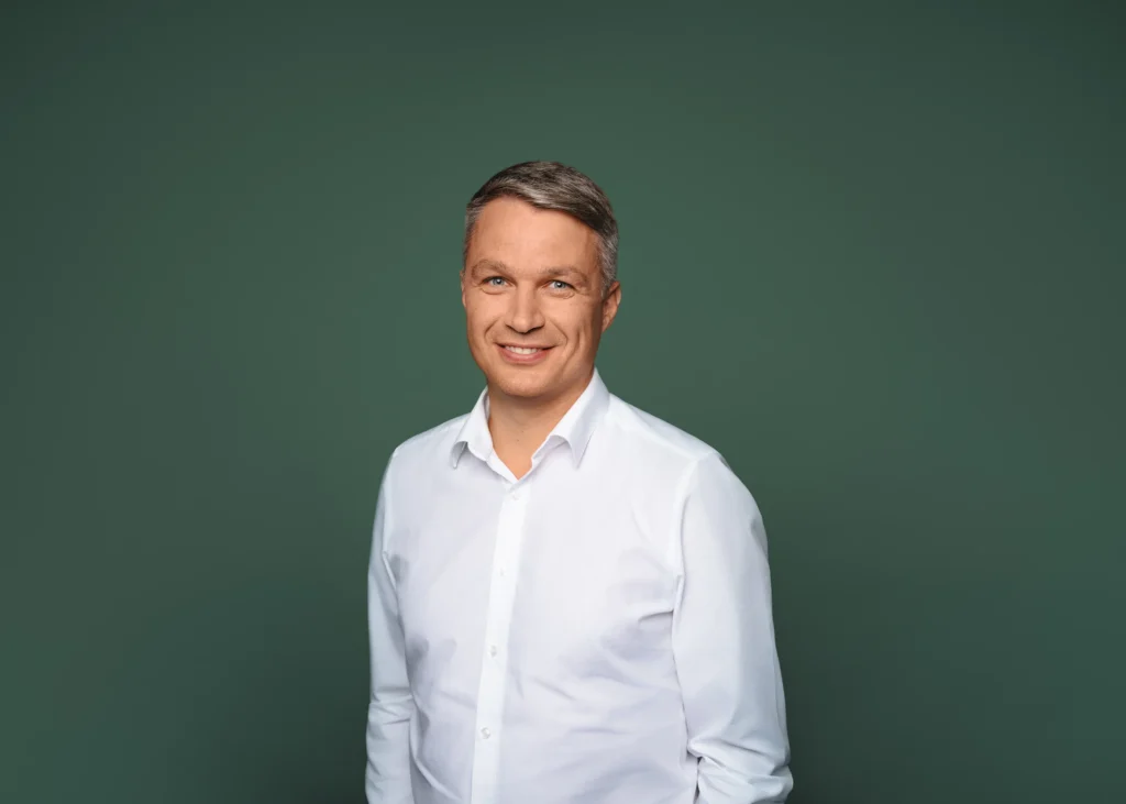 A smiling man with grey hair wearing a white shirt against a solid green background.