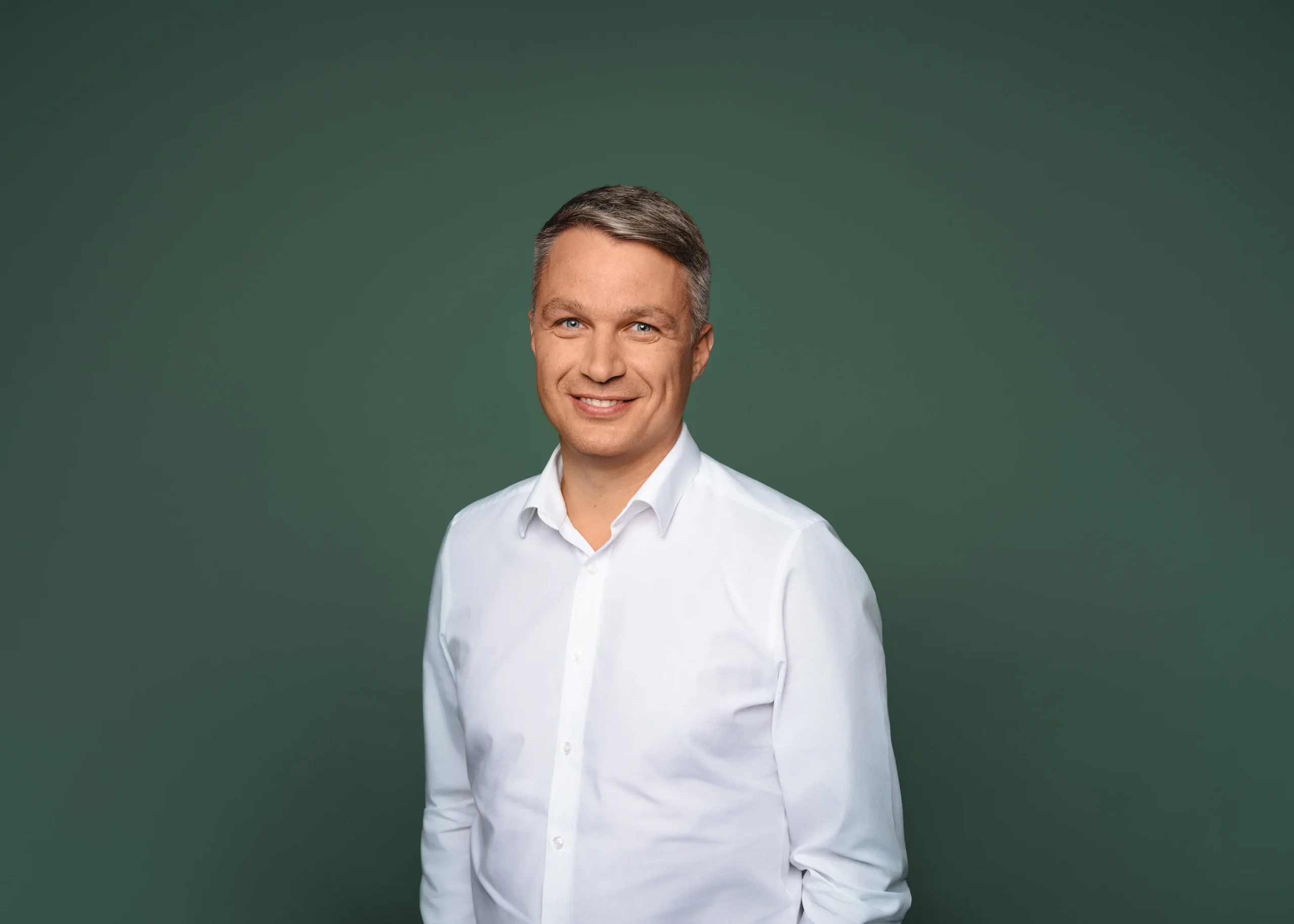 A smiling man with grey hair wearing a white shirt against a solid green background.