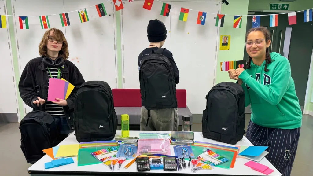 Three students stand behind a table filled with backpacks and various school supply items.
