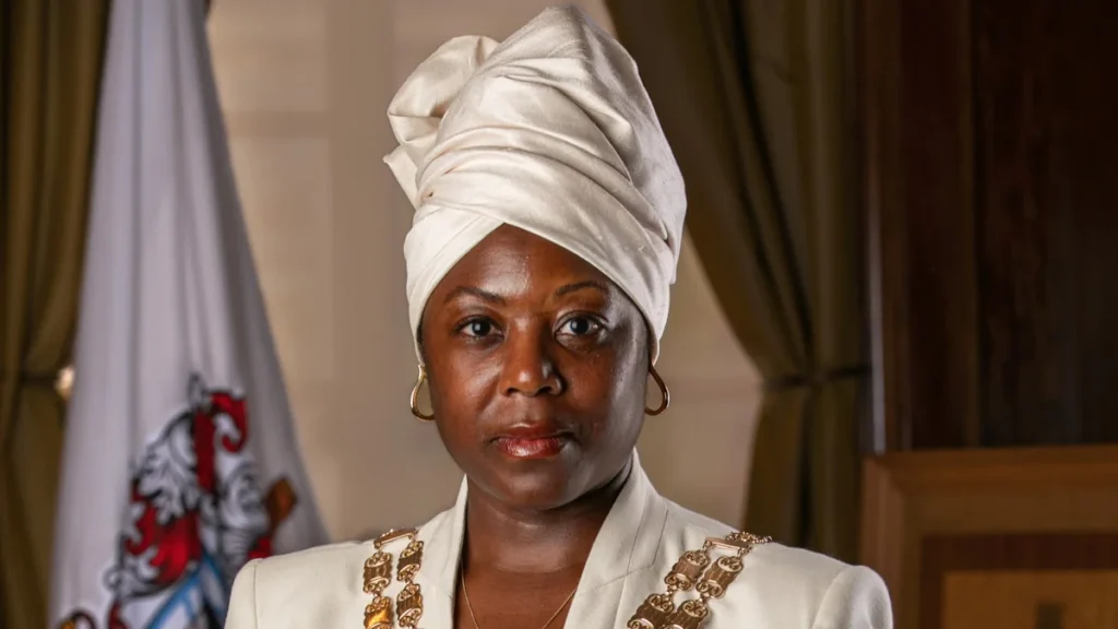 A Black woman wearing a white headwrap and a formal white suit with jewelry.