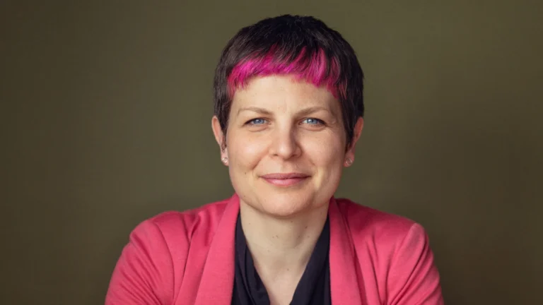 Woman with short hair, pink highlights, and a pink jacket smiling against a green background.