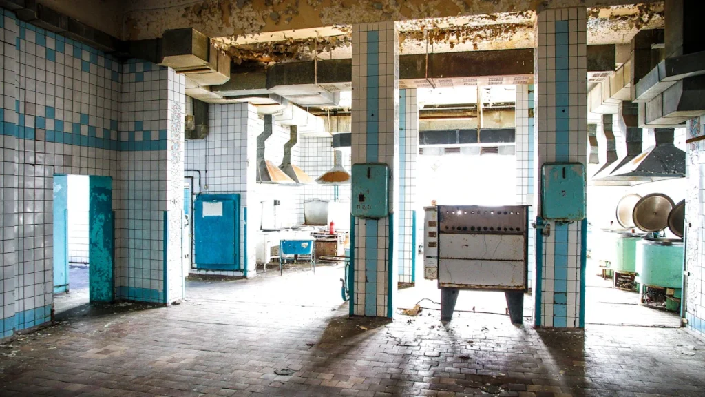 An abandoned and dirty commercial kitchen with peeling paint, old tiles, and industrial ventilation hoods.