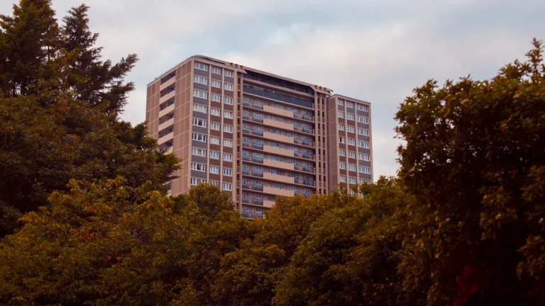 A tall multi-story residential apartment building in London viewed behind a row of green trees.