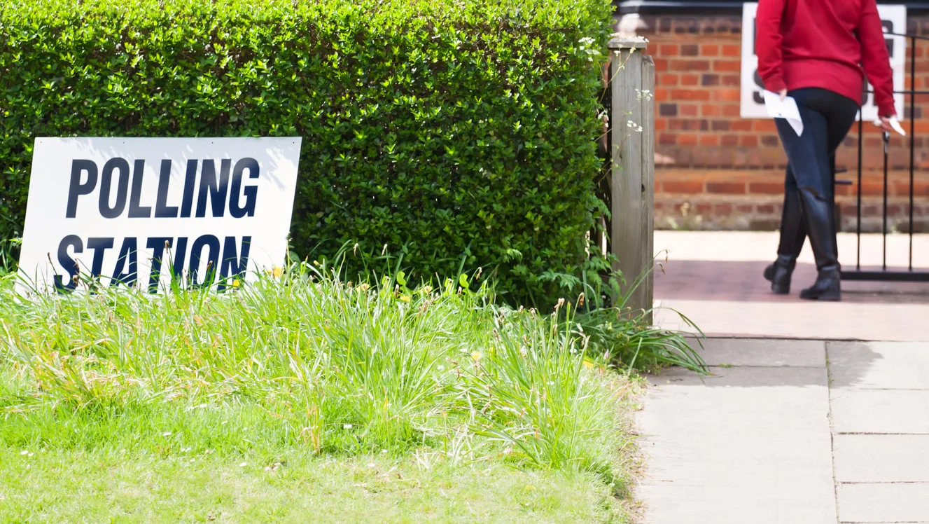 White sign reading 'POLLING STATION' stands in front of a green hedge and tall grass.