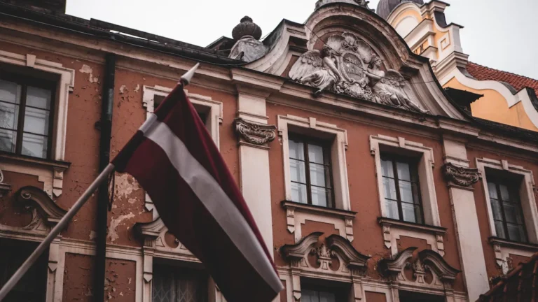 The national flag of Latvia waving in front of a historic, ornate European building in Riga.