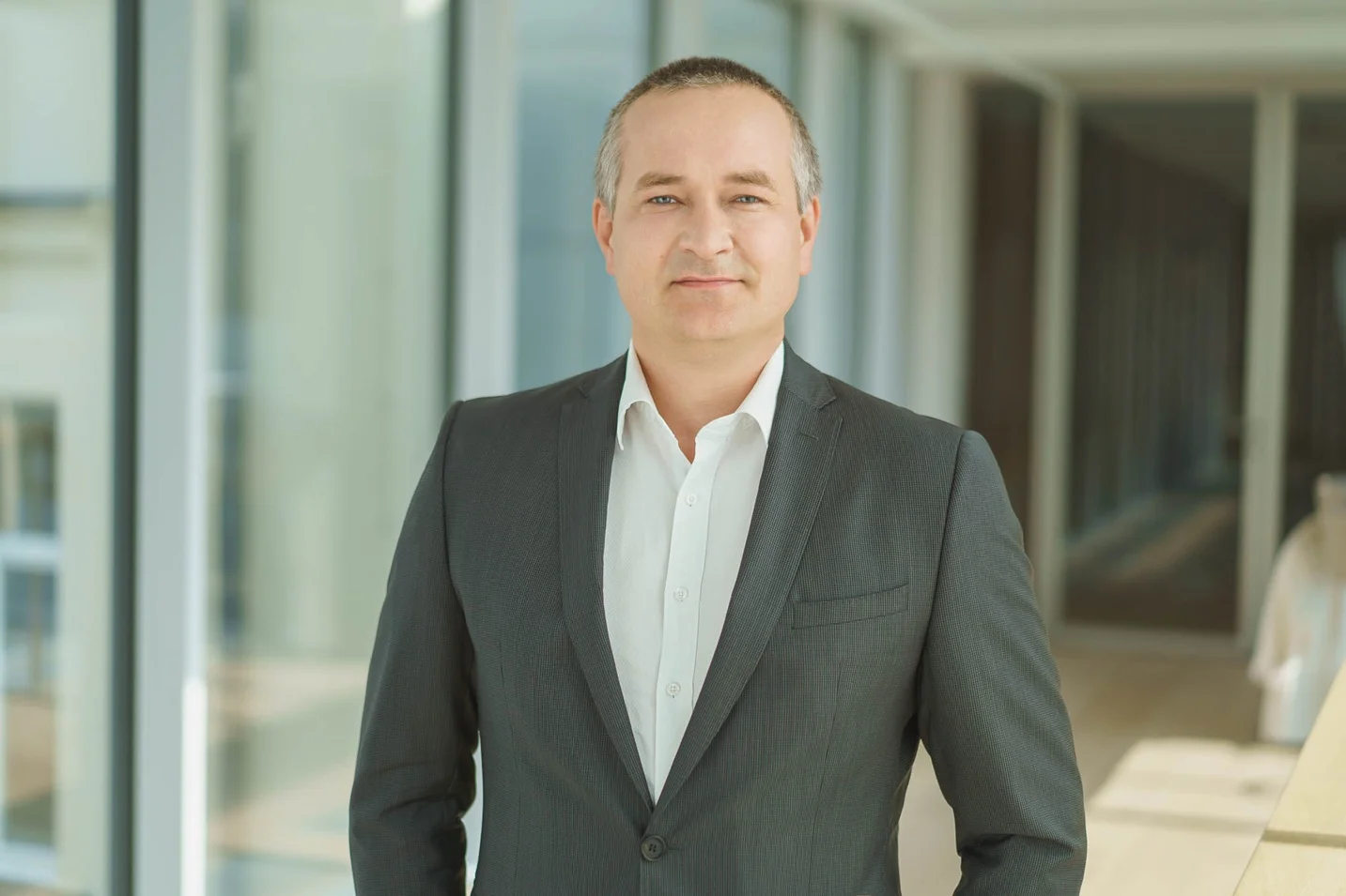 Man in a grey suit and white shirt posing in a bright, modern office hallway.