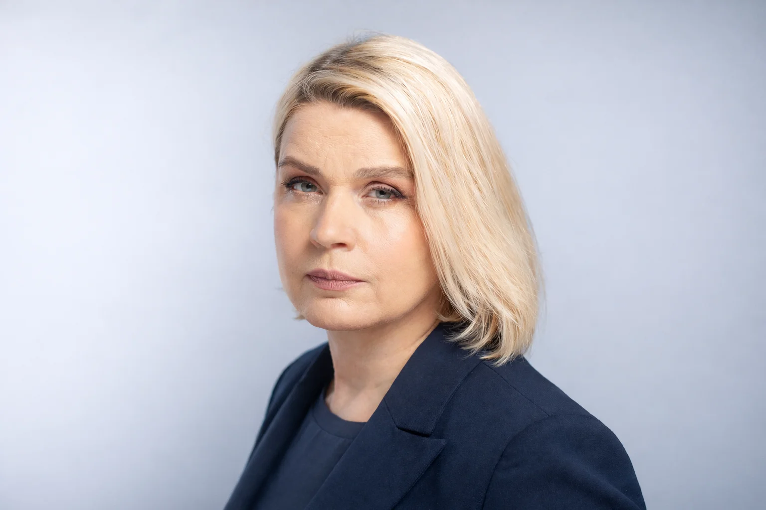 Blonde woman with a serious look wearing a blue blazer against a light studio background.