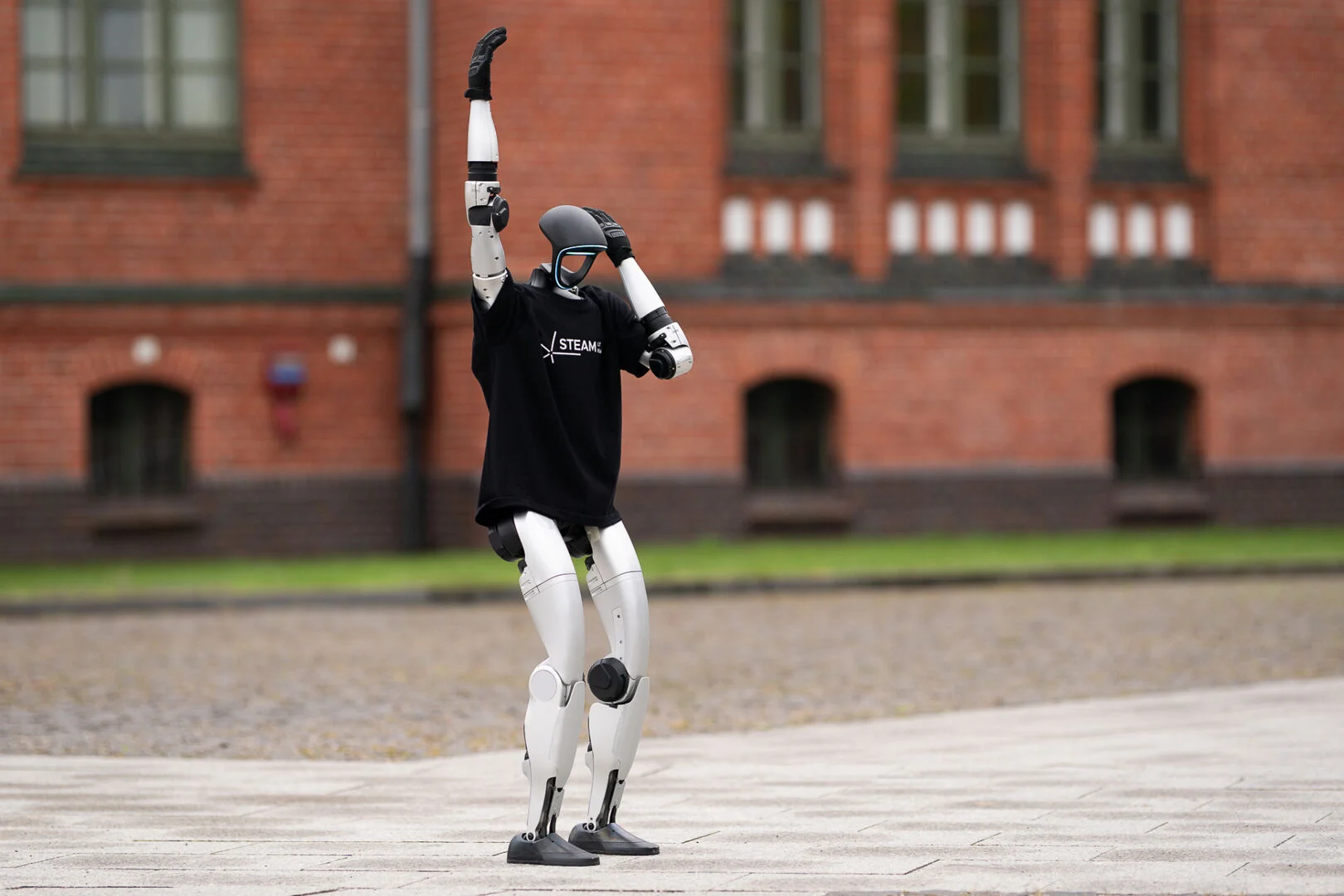 A silver humanoid robot wearing a black t-shirt raises its arm in front of a brick building.