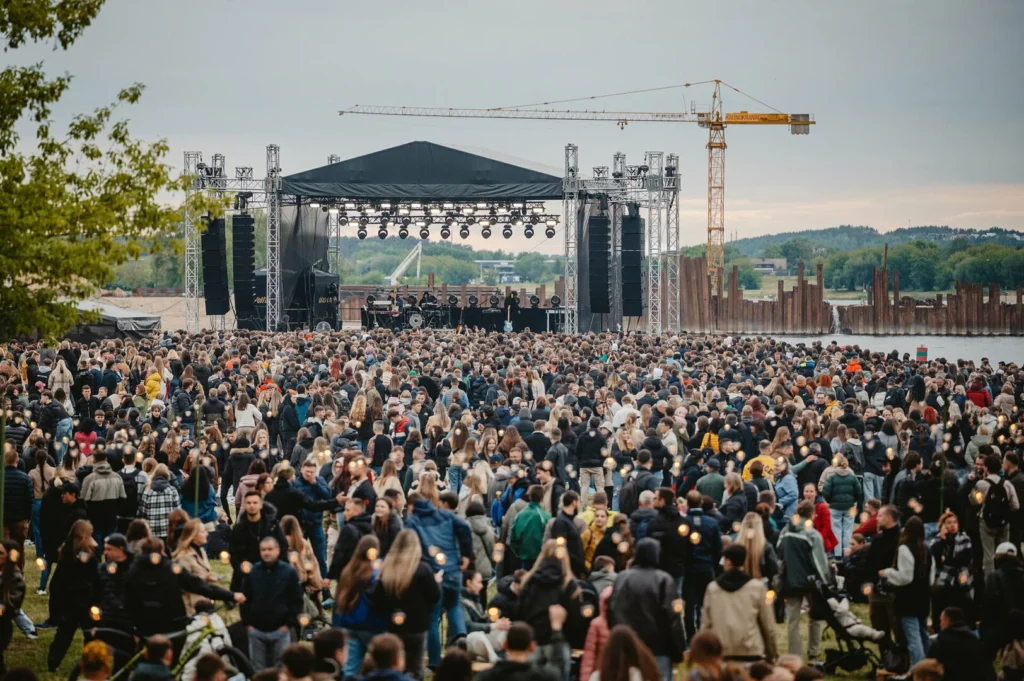 Thousands of people gather before an outdoor concert stage near a riverfront and construction crane.