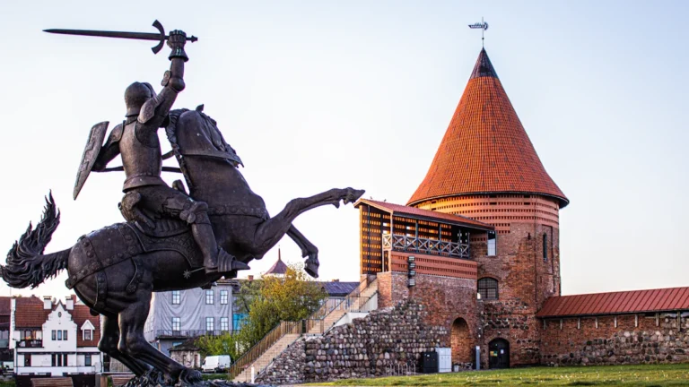 A bronze knight statue stands prominently in front of the red-roofed tower of Kaunas Castle.