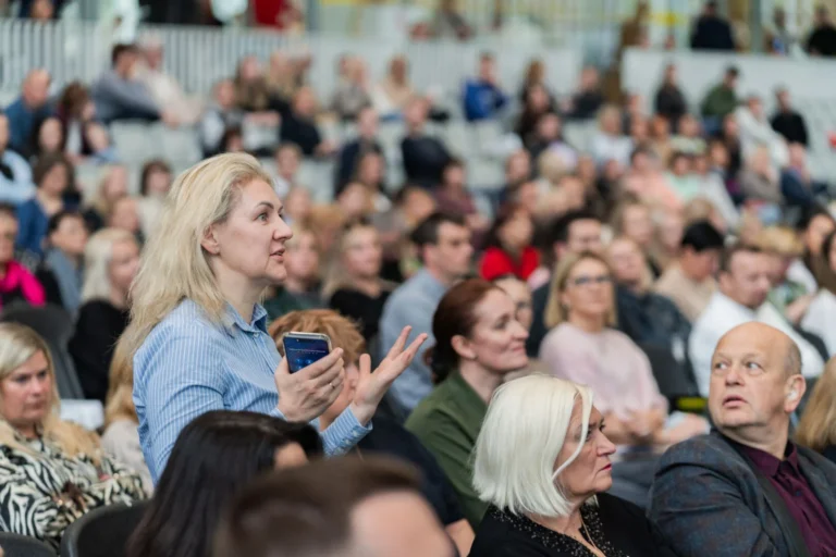 Blonde woman in a blue shirt gestures while speaking to a large, seated audience.