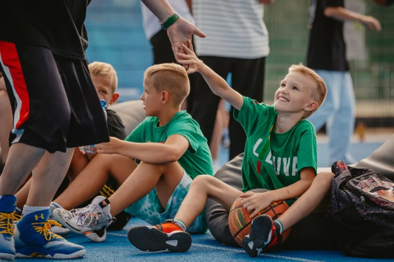 Smiling young boy sitting with a basketball high-fives an adult on an outdoor blue court.