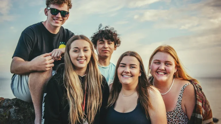 A group of five diverse and smiling teenagers posing together outdoors during the summer.