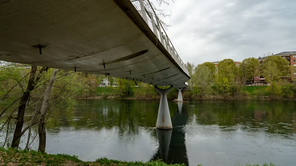 A modern concrete bridge spanning a calm river surrounded by green trees under an overcast sky.
