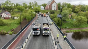 Two heavy trucks and workers on a bridge undergoing renovation with a church background.