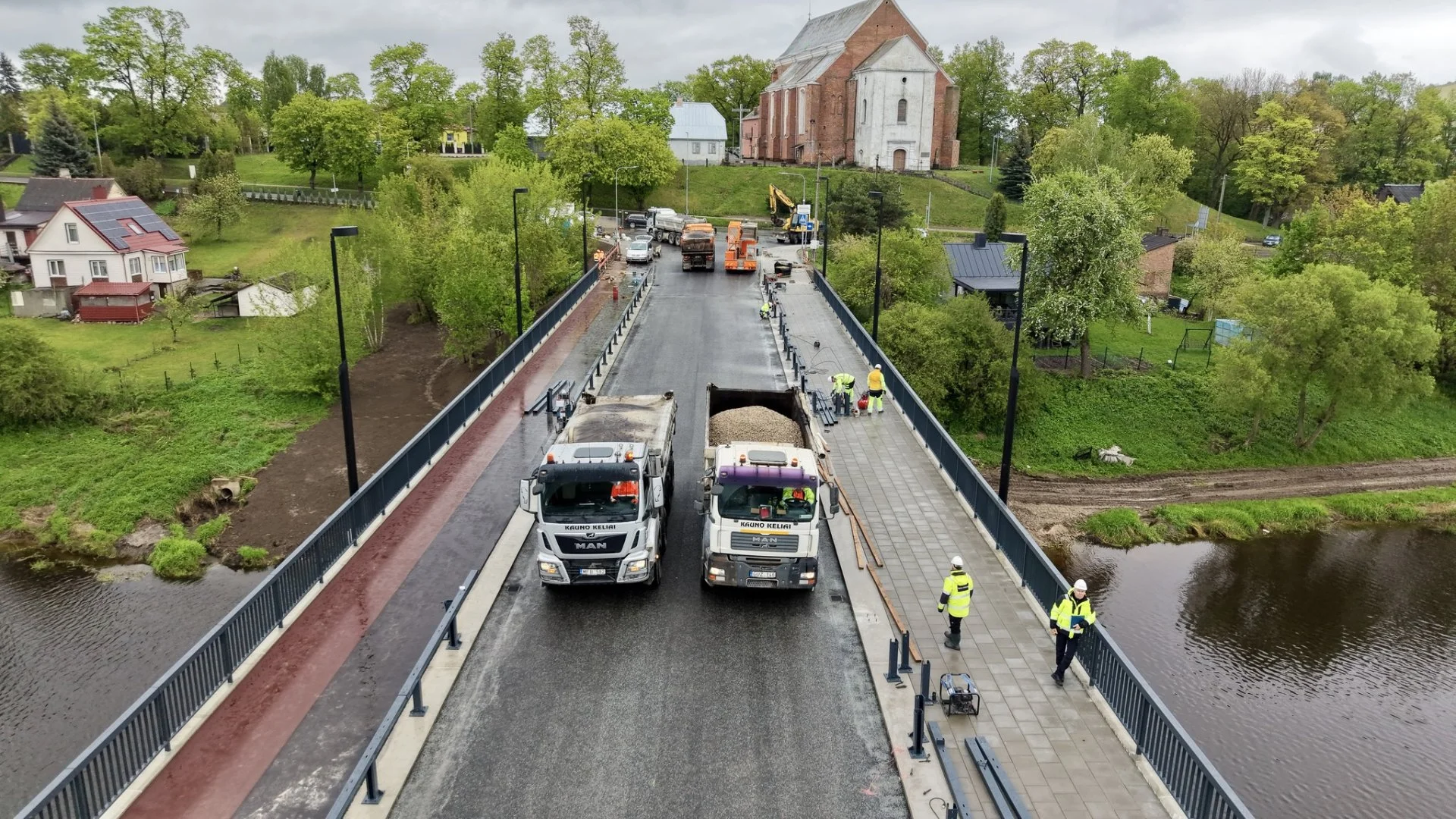 Two heavy trucks and workers on a bridge undergoing renovation with a church background.