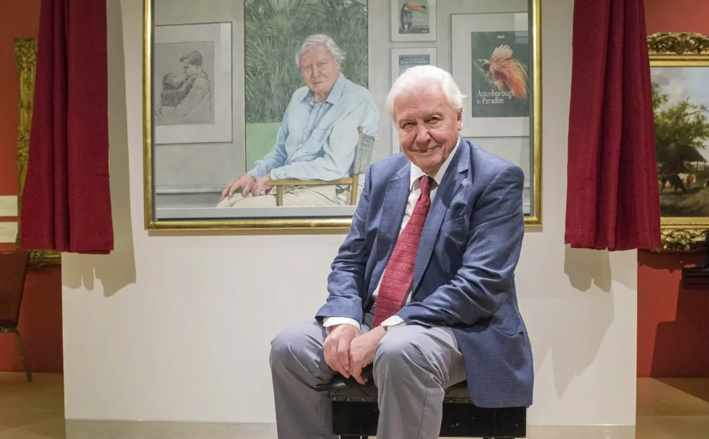 Sir David Attenborough sits in front of a large portrait of himself in an art gallery.
