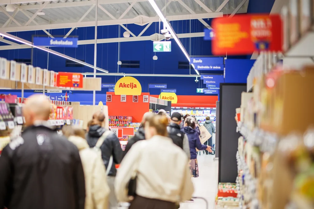 Crowded grocery store aisle with shoppers walking past yellow promotion signs and various product shelves.