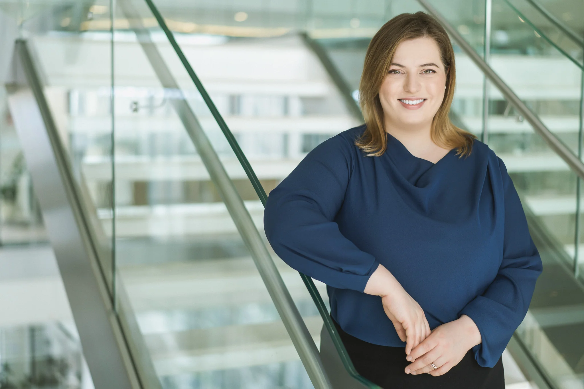 Smiling woman wearing a blue blouse, leaning against a glass staircase railing in a bright office.