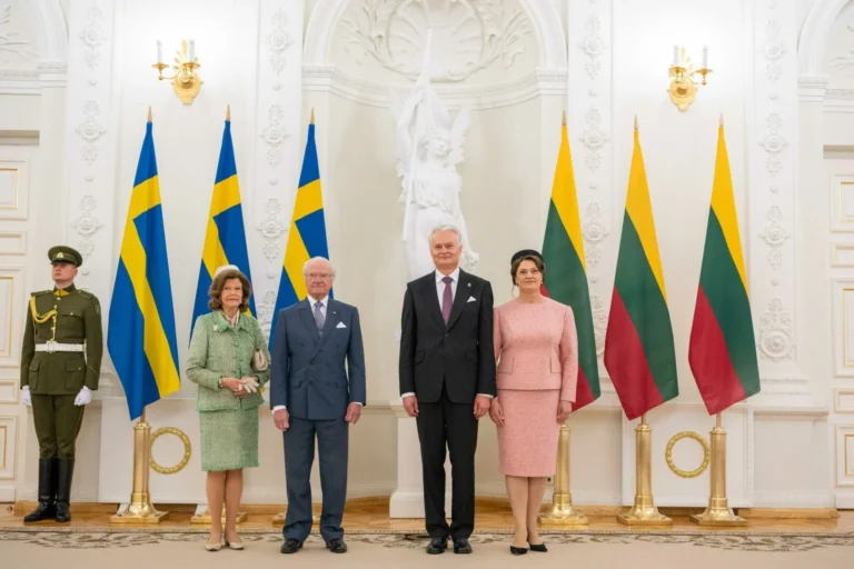 Swedish royals and Lithuanian presidential couple stand formally between national flags in a white hall.