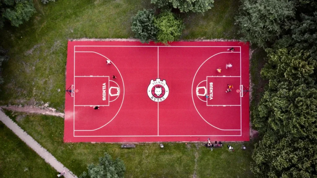 Top-down aerial view of a red basketball court in Vilnius, Lithuania, bordered by lush green trees.