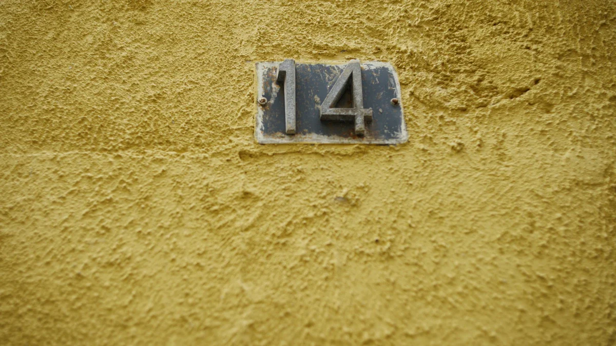 A metal house number 14 mounted on a textured yellow stucco exterior wall in an urban setting.