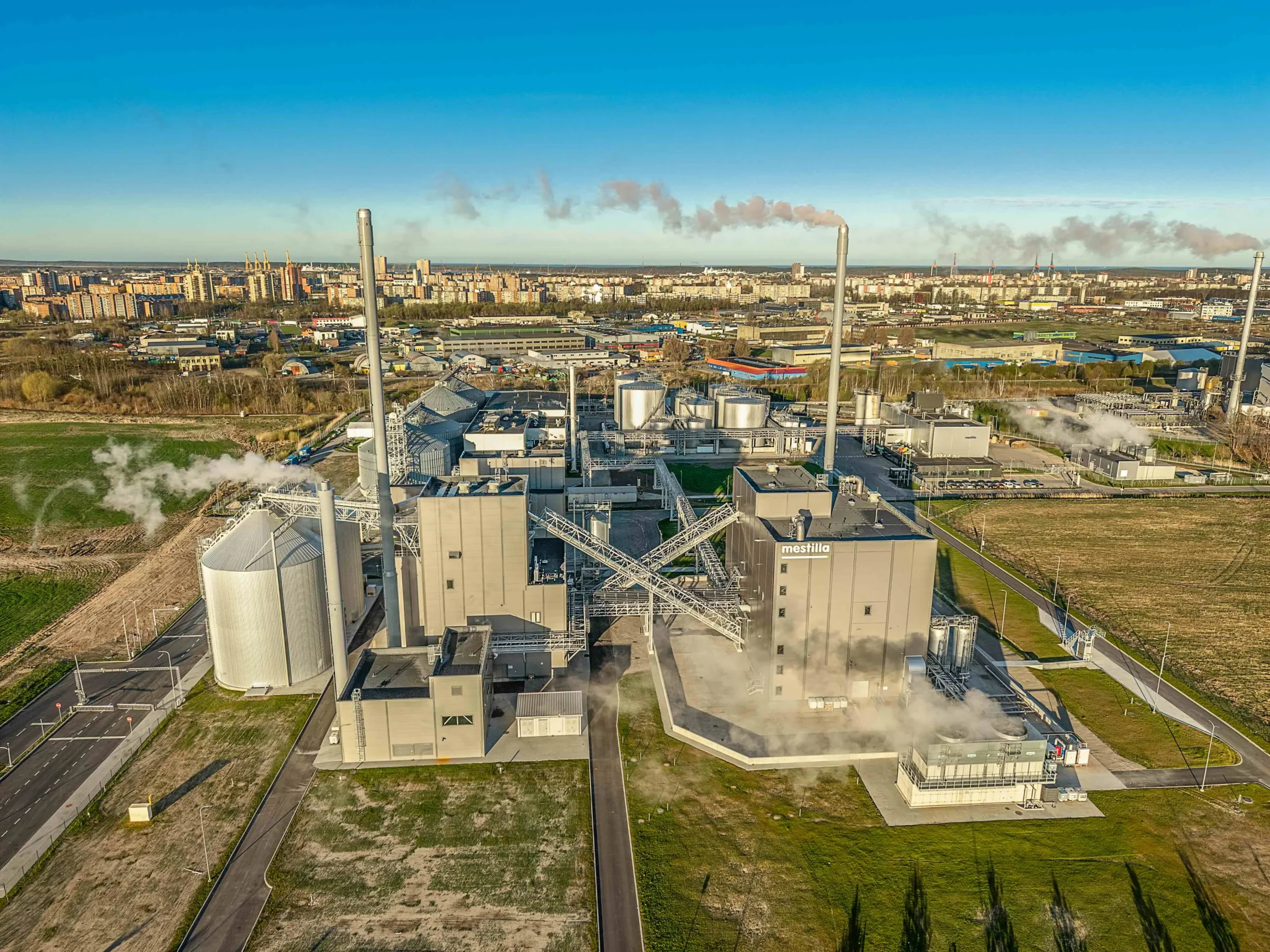 Aerial view of a large industrial plant with tall smokestacks emitting steam near a city.