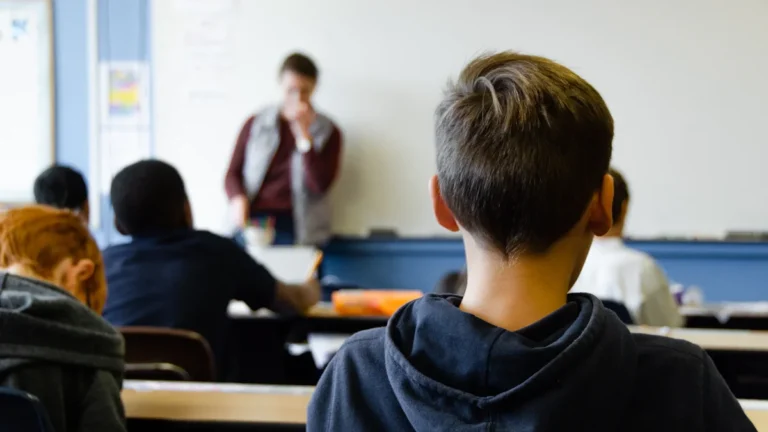 A young student in a dark hoodie sits at a desk in a classroom setting.